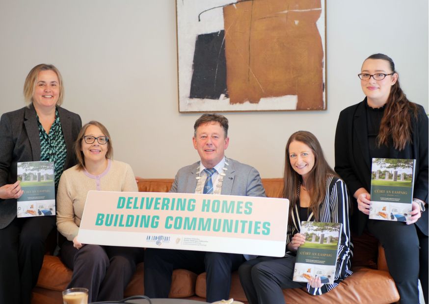 Five people sitting around couch in sitting room of new house holding corribiard sign